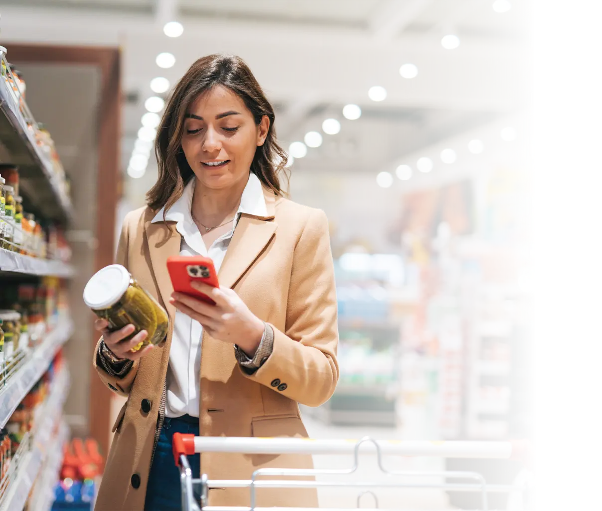 A woman shopping, while holding her phone and jar of pickle 